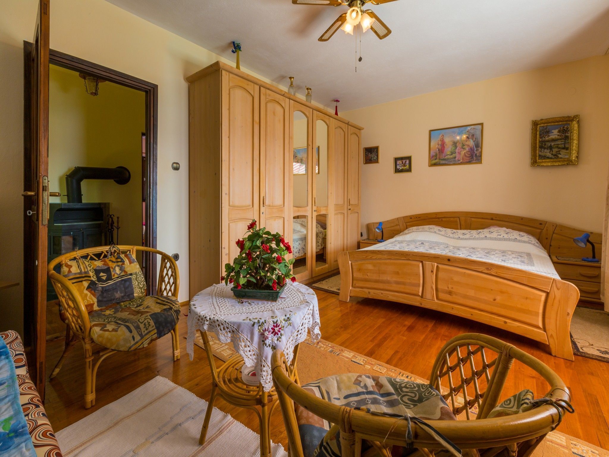 Ein Schlafzimmer mit einem hölzernen Bett, einem Holzschrank, einem Korbstuhl, einem kleinen Tisch mit einer Spitzenwäsche und einem Deckenventilator.