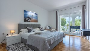 A modern bedroom with a large bed, a bedside table with a lamp, a view of the outdoors through a sliding glass door, and a painting on the wall. - Apartment Villa Zdenka (Crikvenica)