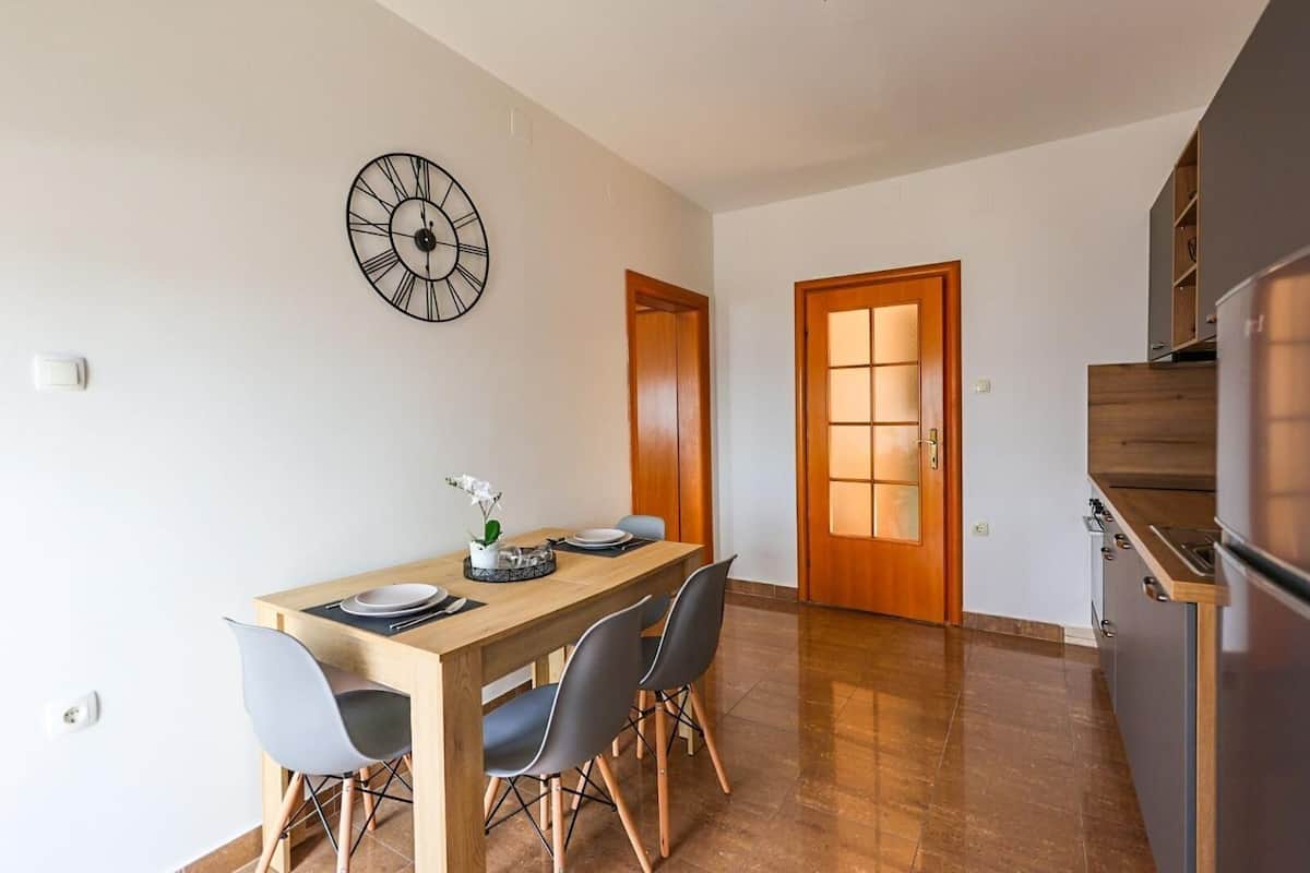 A dining area with a wooden table, four chairs, a clock on the wall, and a kitchen with wooden cabinets and a refrigerator.