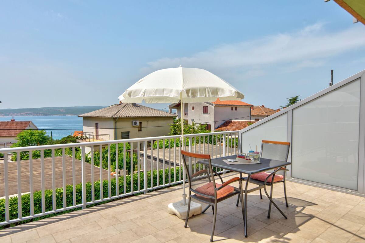 A balcony with a table and chairs, a white umbrella, and a view of the sea and buildings.
