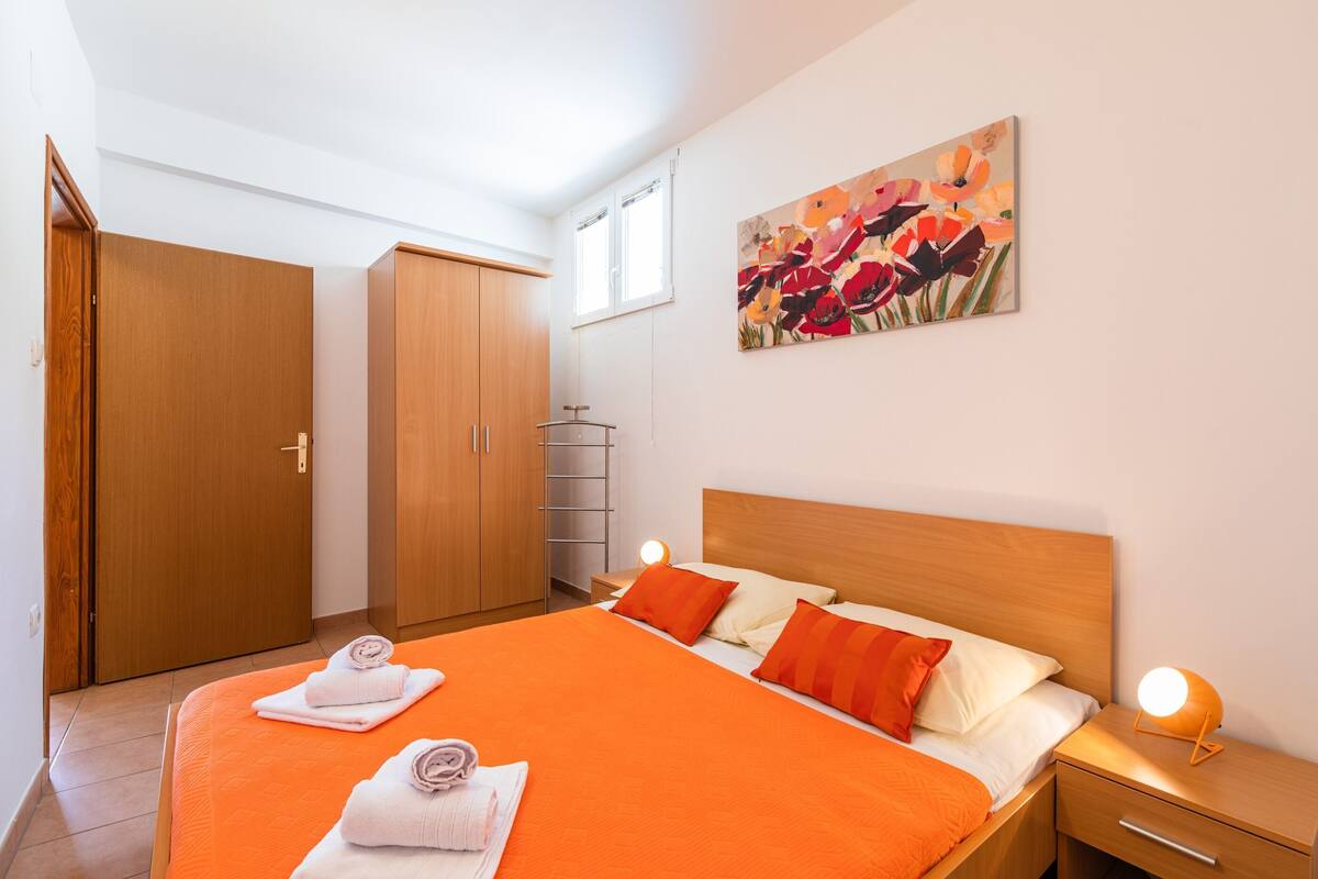 A hotel room with a large bed, orange bedding, a wooden headboard, a bedside table with a lamp, a painting on the wall, and a wardrobe.