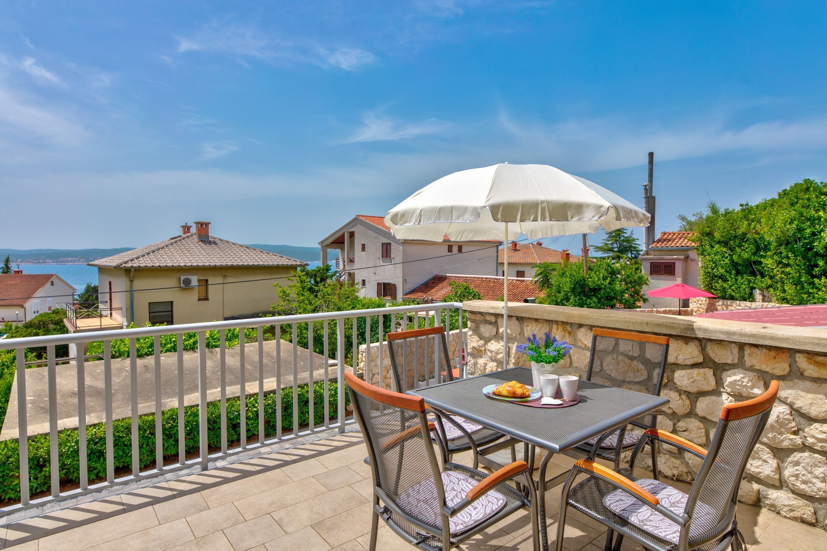 Ein Balkon mit Tisch und Stühlen, einem weißen Sonnenschirm und Blick auf Häuser und Grünflächen.