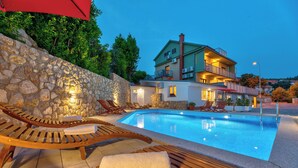 Area piscina con lettini, un muro in pietra e un edificio con balconi e illuminazione esterna.