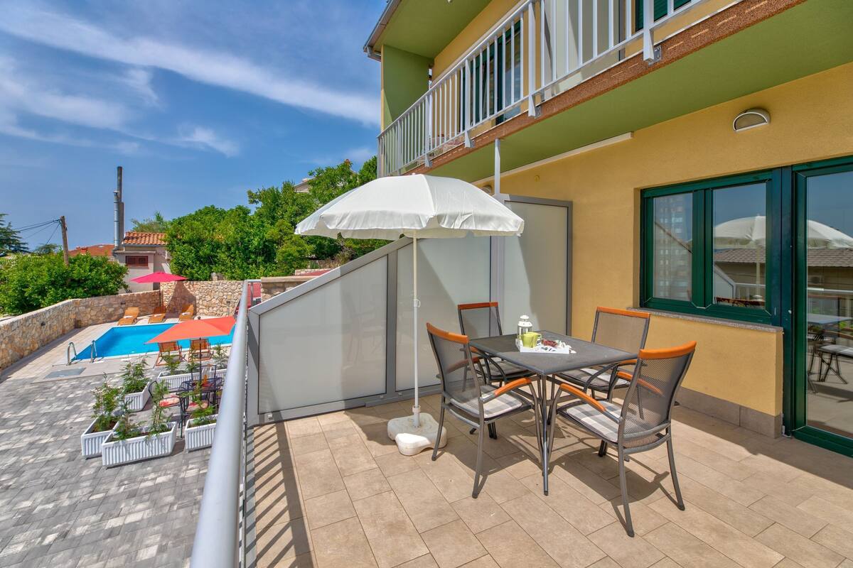 A balcony with a table and chairs, a white umbrella, and a swimming pool.