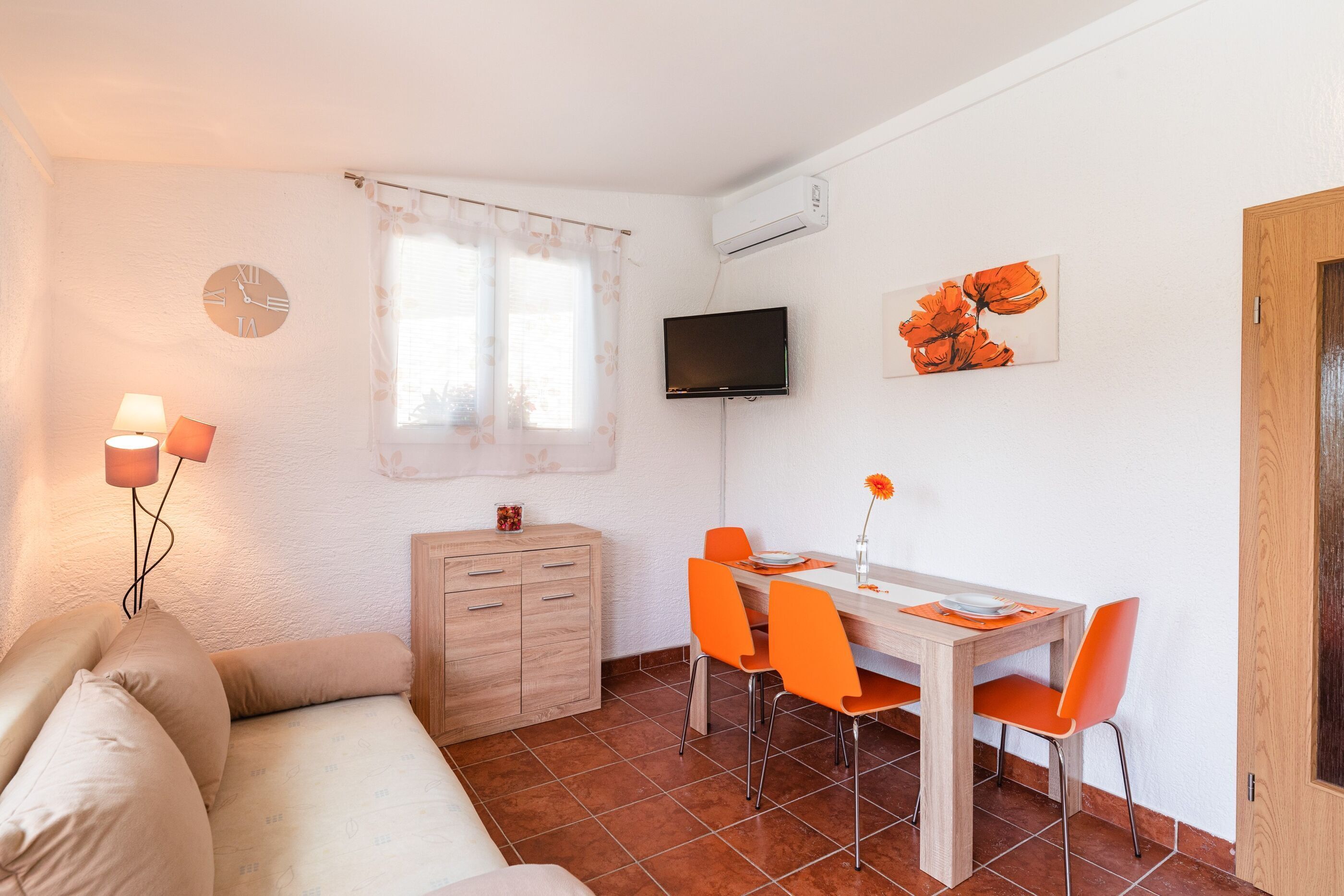 A small, well-lit room with a sofa, a wooden cabinet, a dining table with orange chairs, a TV mounted on the wall, and a window with curtains.