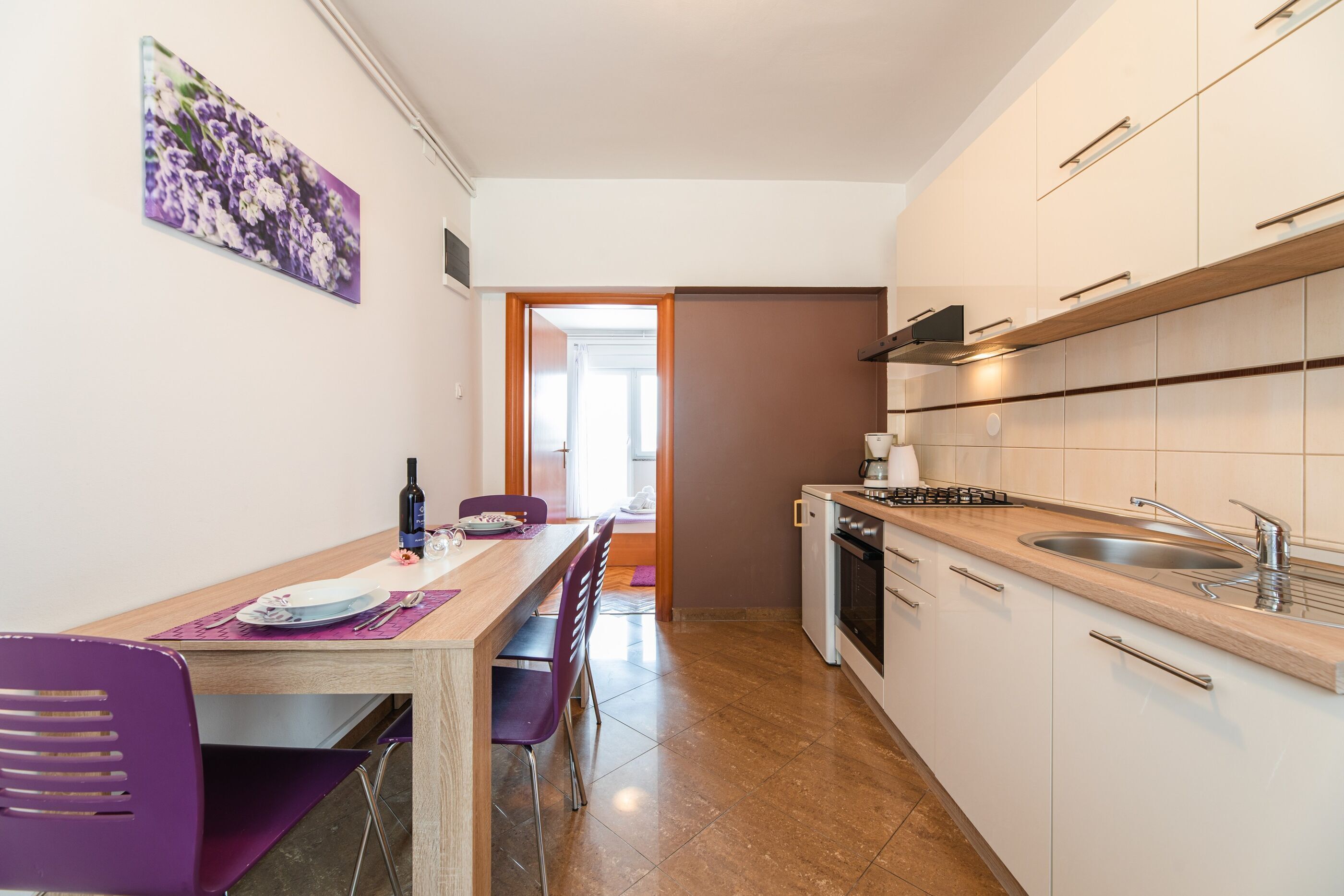 Eine kleine Küche mit Essbereich, mit einem Holztisch, violetten Stühlen und Blick in den Speisesaal.