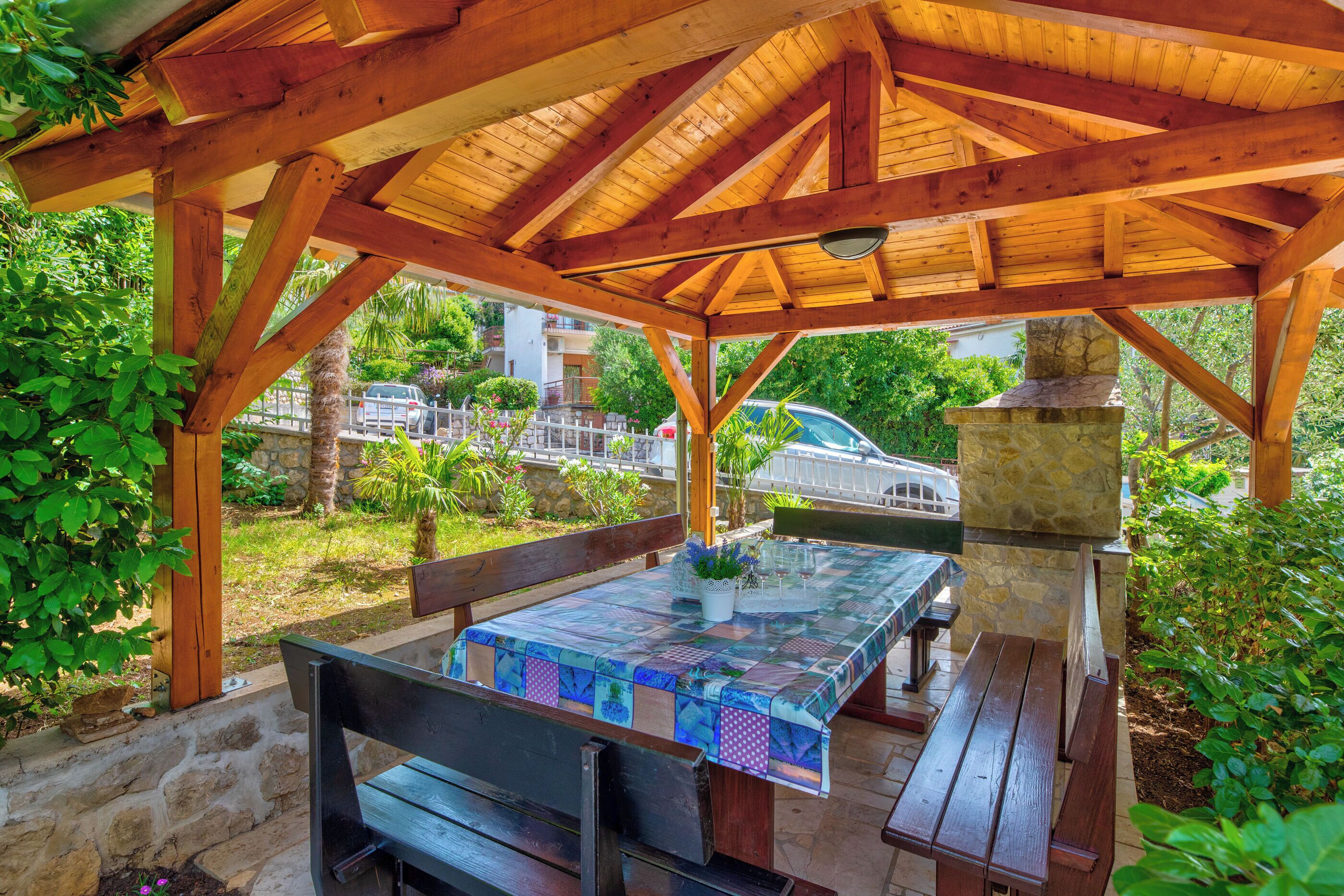 A wooden gazebo with a table and benches, surrounded by greenery.
