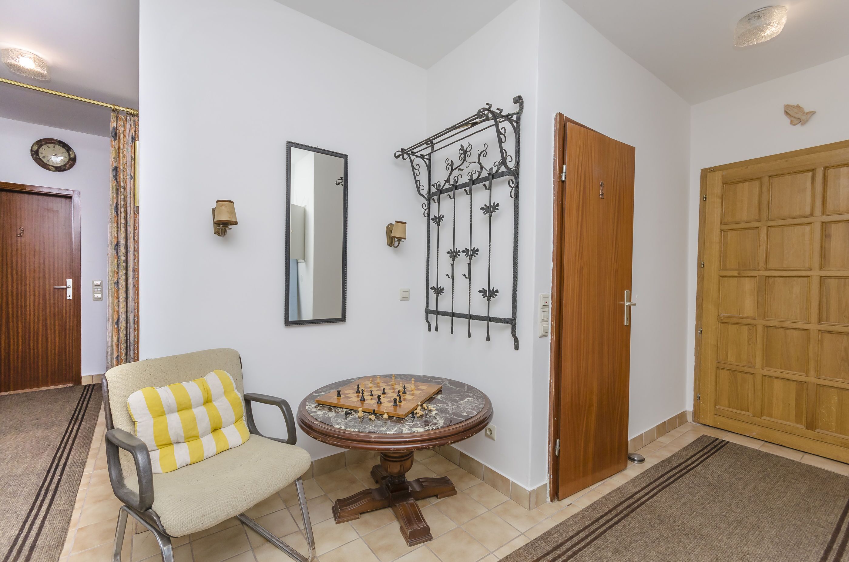 A room with a wooden door, a mirror, a chessboard, a chair with a striped cushion, and a wall-mounted light fixture.