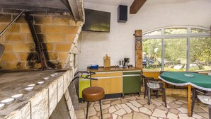 Une cuisine avec un four en briques, un comptoir en bois, un tabouret vert et une table recouverte d’une nappe verte.
