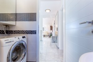 A modern laundry room with a front-loading white washer and dryer, a tiled backsplash, and a hallway leading to a living area with a sofa and a coffee table. - Apartment Dario (Crikvenica)