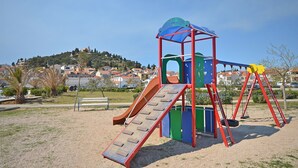 A playground with a slide and swings, set against a backdrop of houses and a hill. - Apartment and Room Ilija (Tribunj)