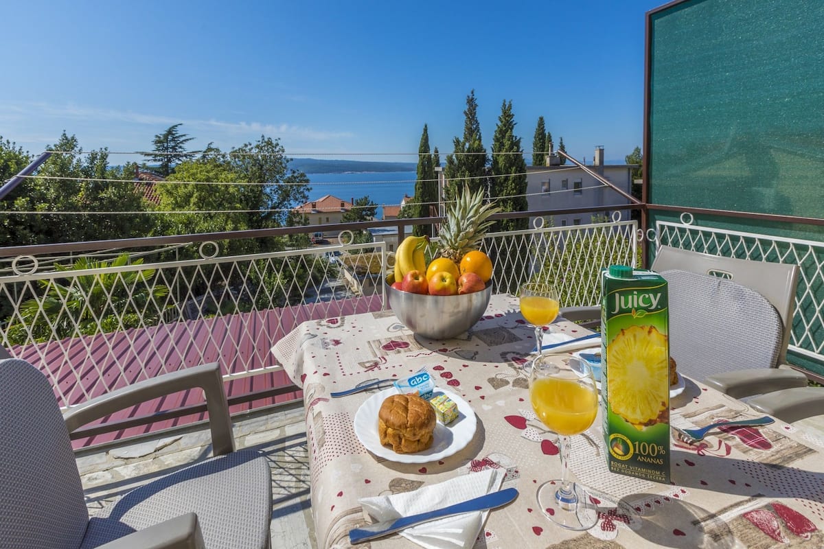 A balcony with a table set for breakfast, including a bowl of fruit, a glass of juice, and a bottle of juice.