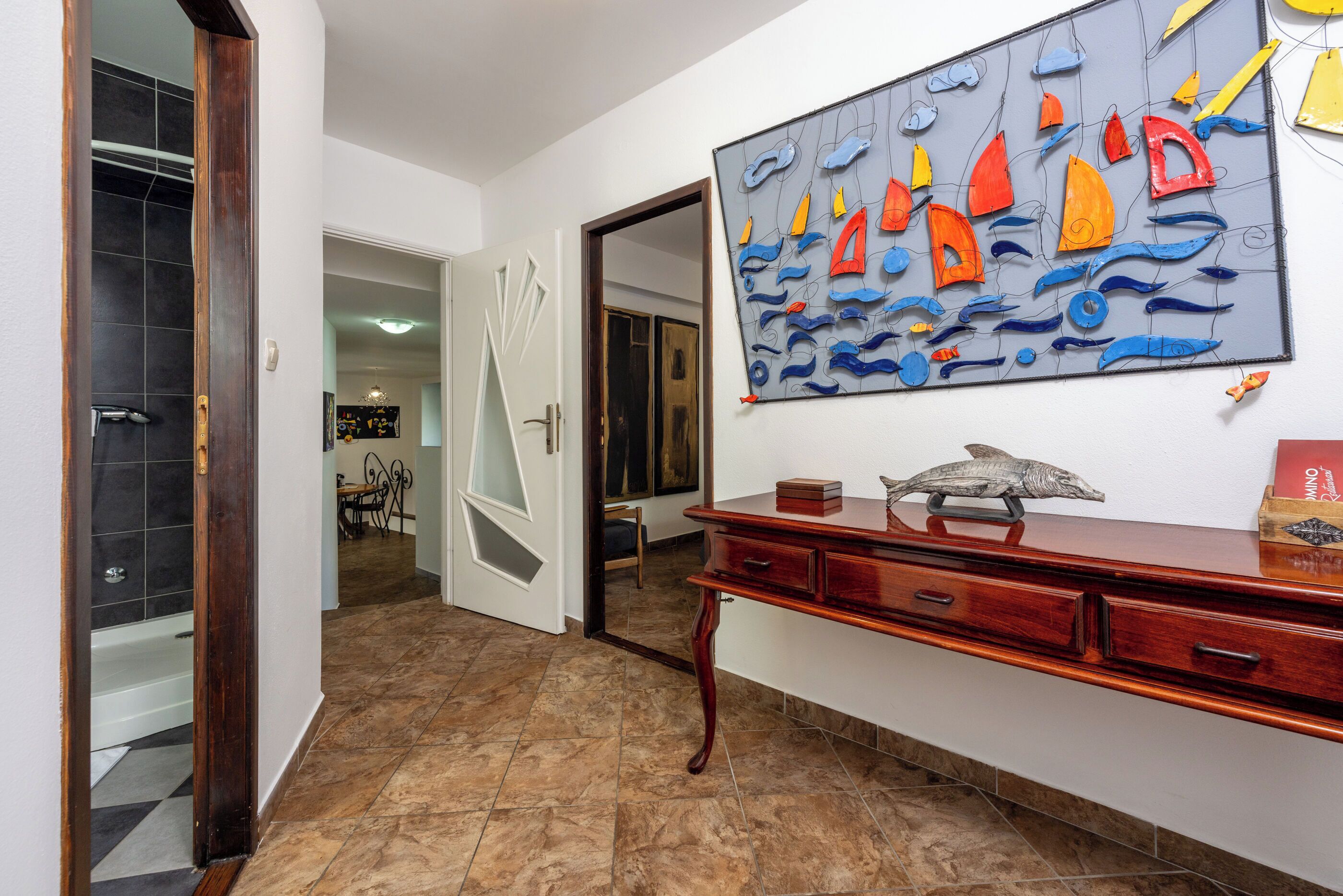 A hallway with a wooden door, a tiled floor, a wooden dresser, and a wall-mounted abstract art piece.