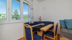 A small room with a wooden table and blue upholstered chairs, a sofa with patterned cushions, and a window with a view of greenery. - Apartment Regan (Crikvenica)