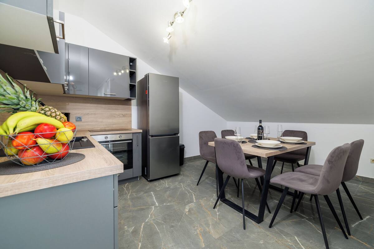 A modern kitchen with a dining area, featuring a refrigerator, oven, and a fruit basket on the counter.
