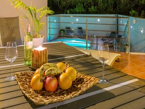 Une table avec un panier de fruits, deux verres à vin et une bougie. Au fond, on aperçoit un espace piscine avec des chaises longues.