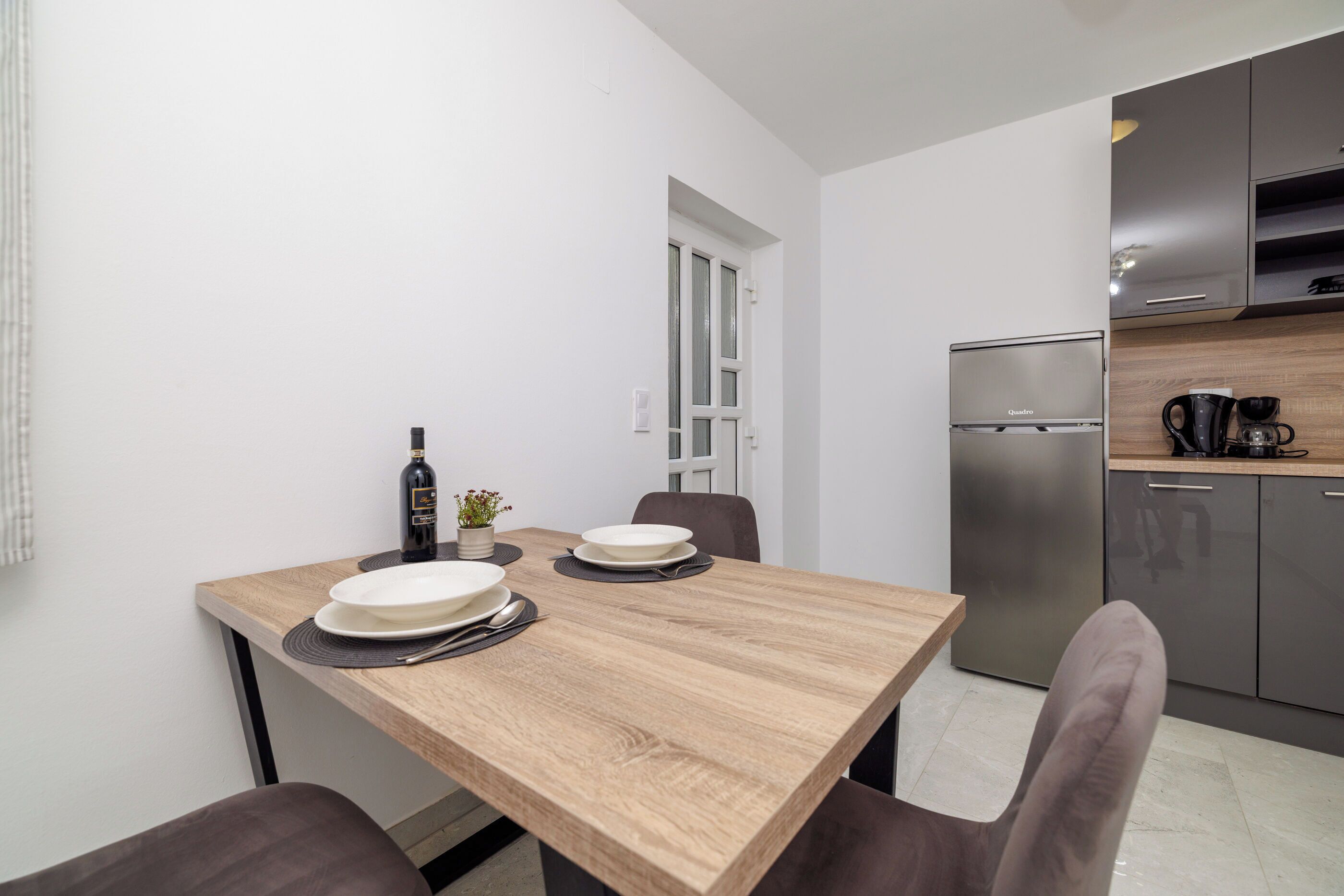 A compact kitchen with a wooden table set for dining, a refrigerator, and a coffee machine.