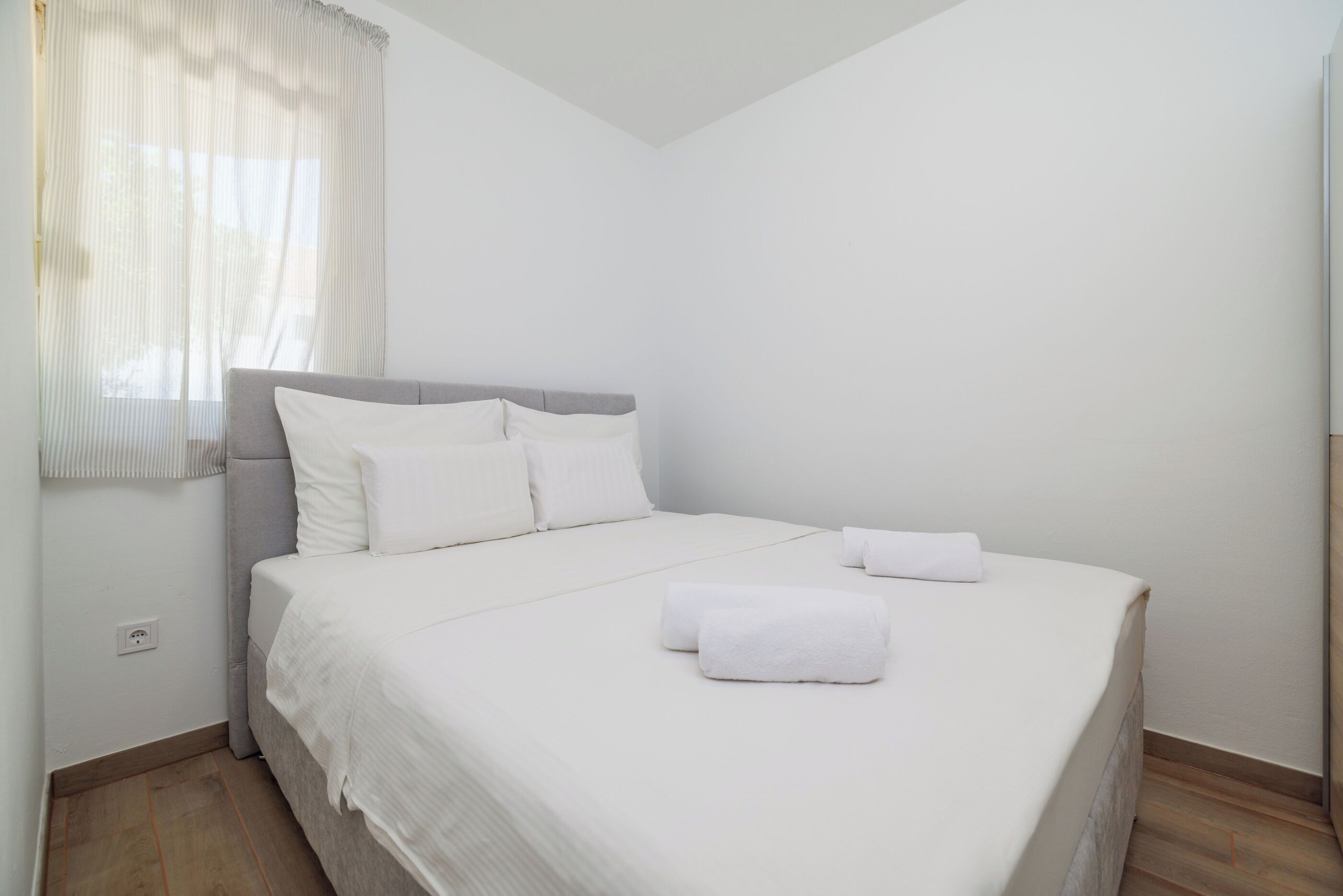 A neatly made bed with white linens and pillows, a window with sheer curtains, and a light switch on the wall.