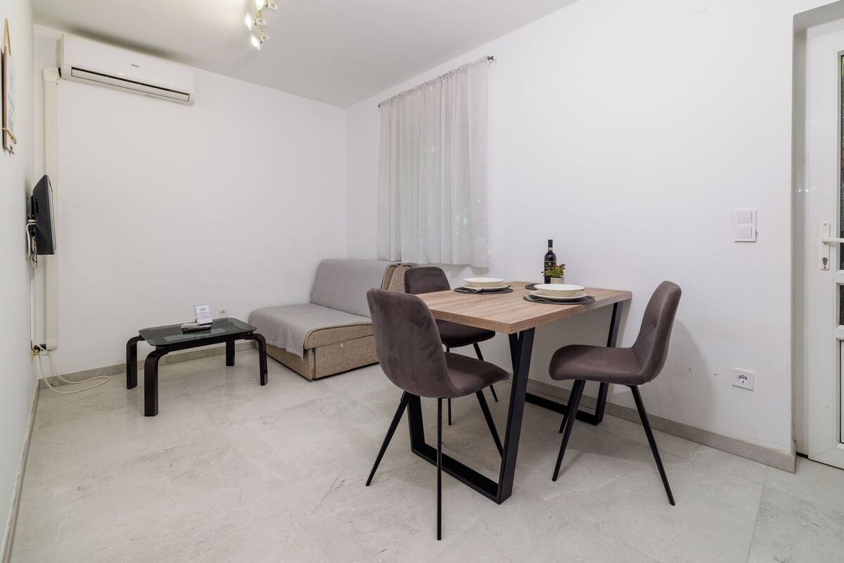A small, modern living space with a dining table, two chairs, a sofa, a small side table, and a TV.