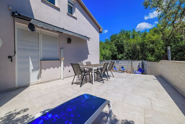 Une terrasse avec une table et des chaises, une piscine et un bâtiment doté de volets.