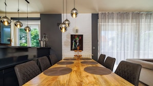 A dining area with a long wooden table, multiple chairs, pendant lights, a painting on the wall, and a window with curtains. - Apartments Alin (Dramalj)