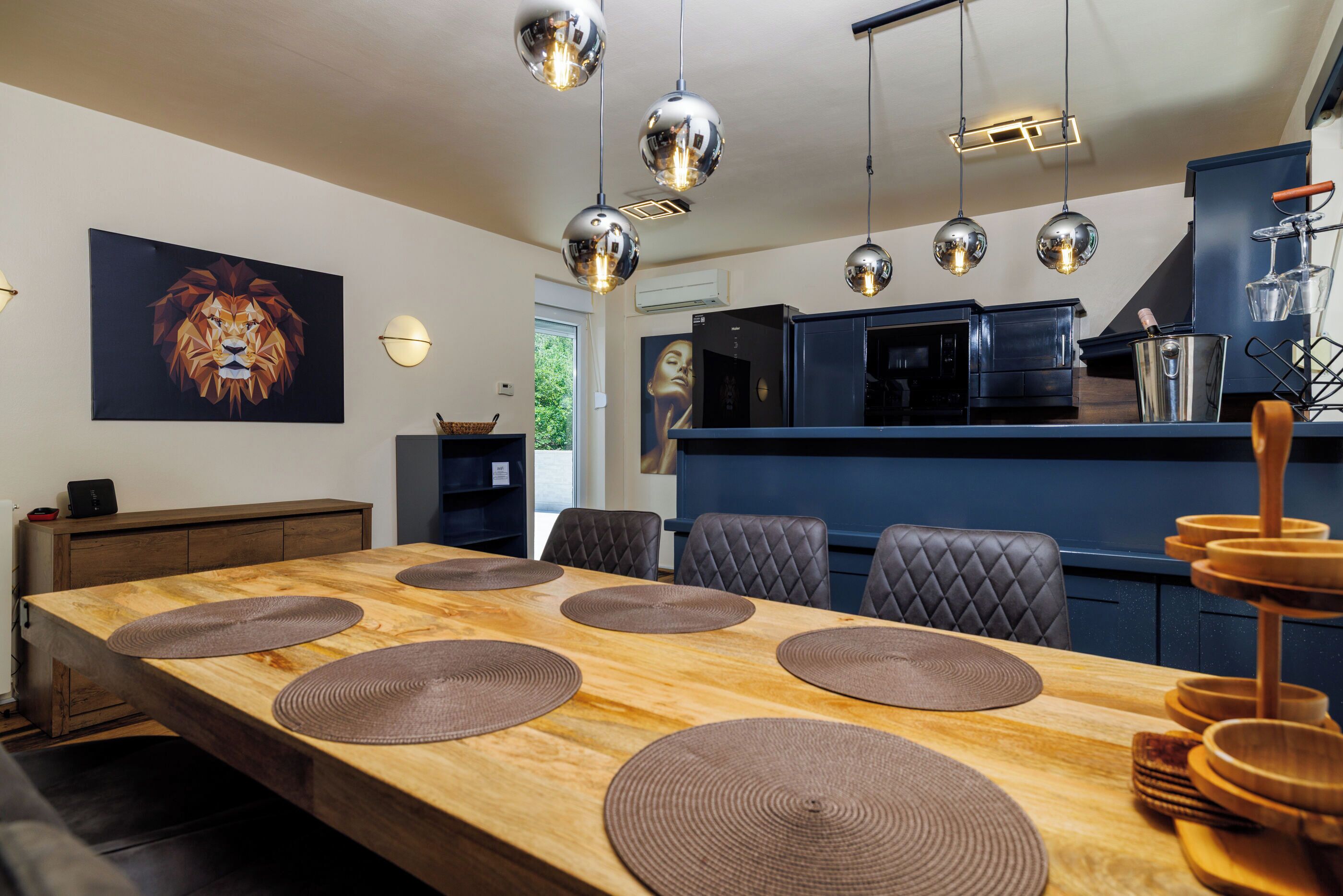 A modern kitchen with a dining area, featuring a long wooden table with placemats, pendant lights, and a large framed artwork on the wall.