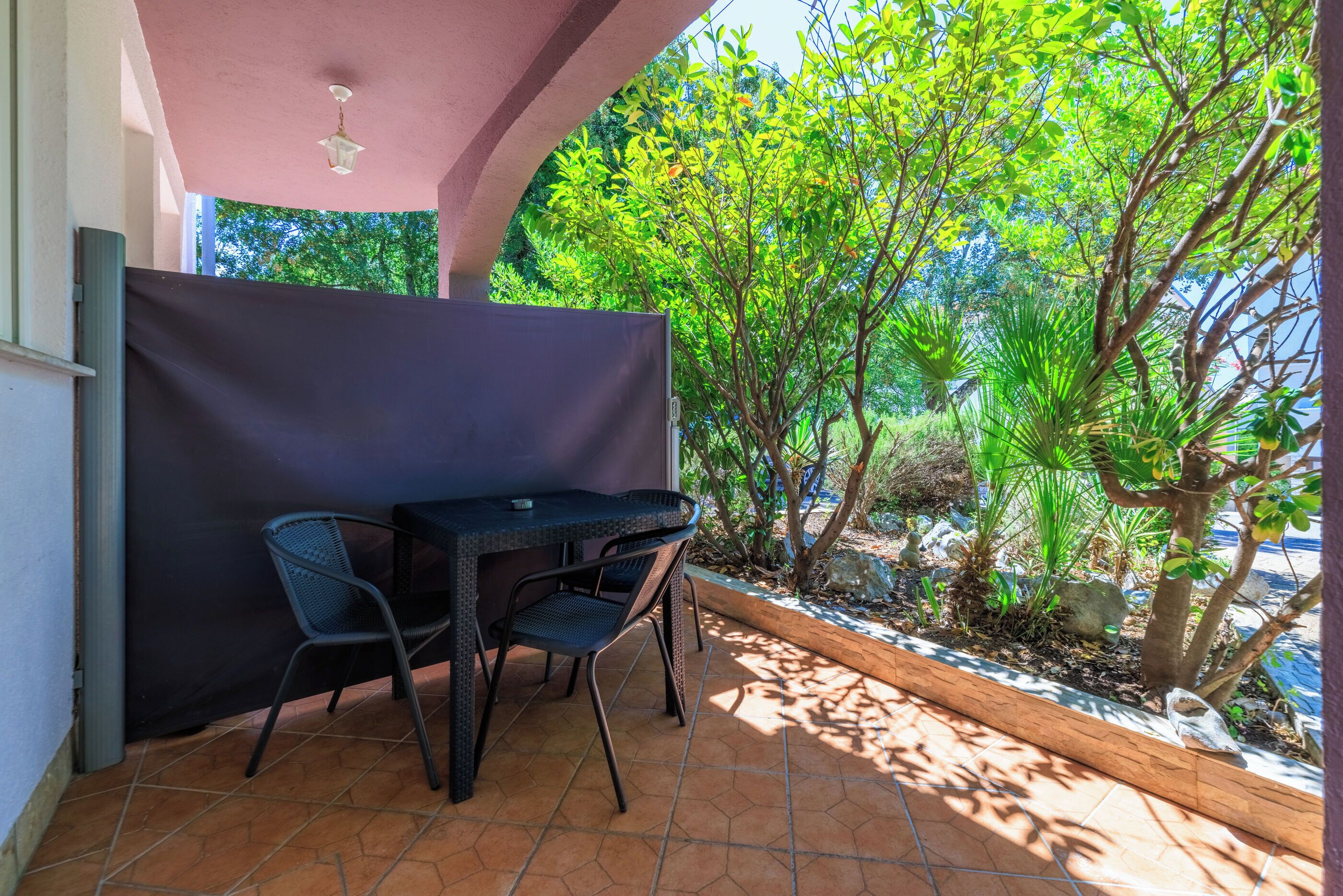 En overdækket terrasse med et bord og stole, omgivet af grønt og udsigt til træer.