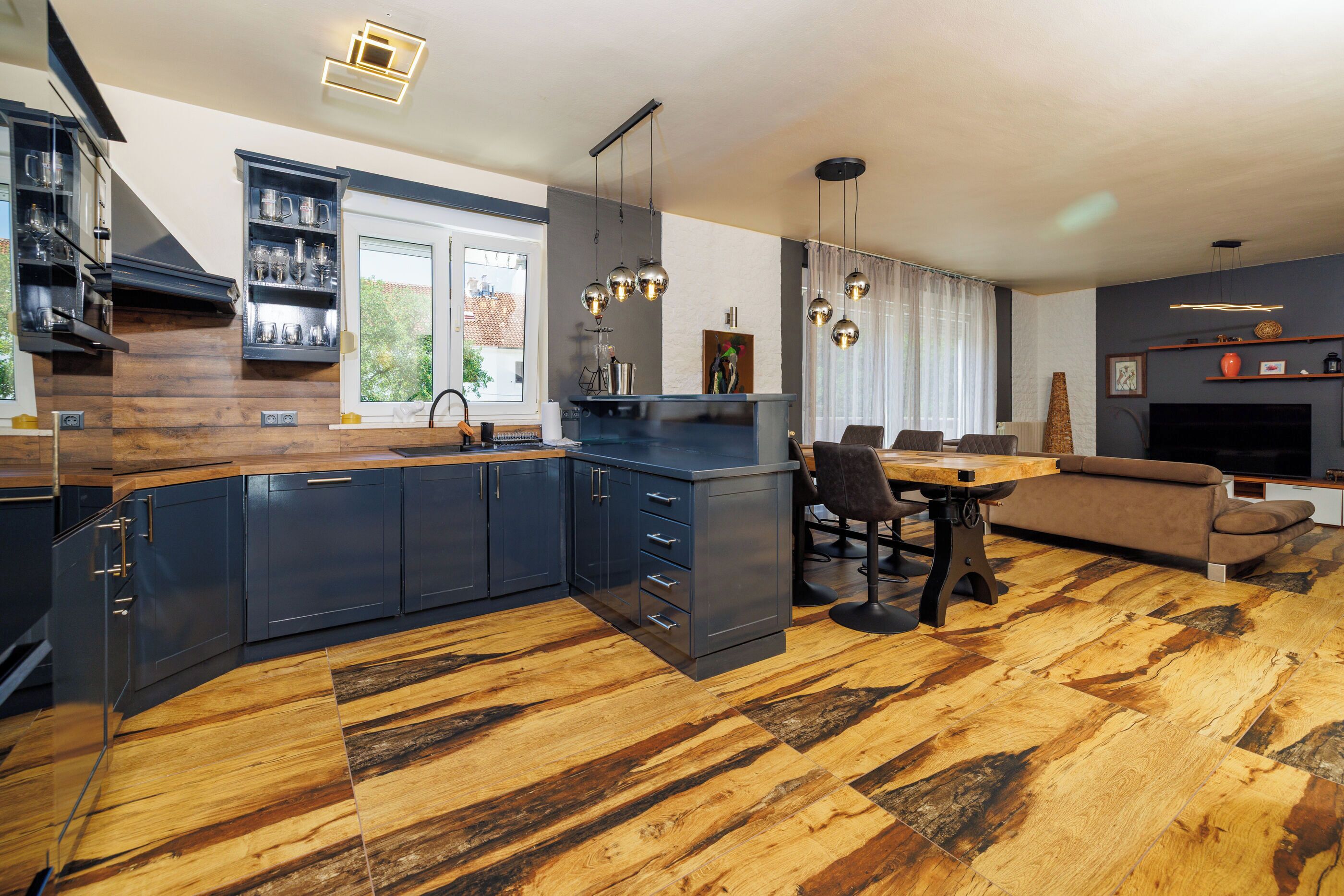 A modern kitchen with dark cabinetry, a dining area with a table and chairs, and a living room with a sofa and a fireplace.