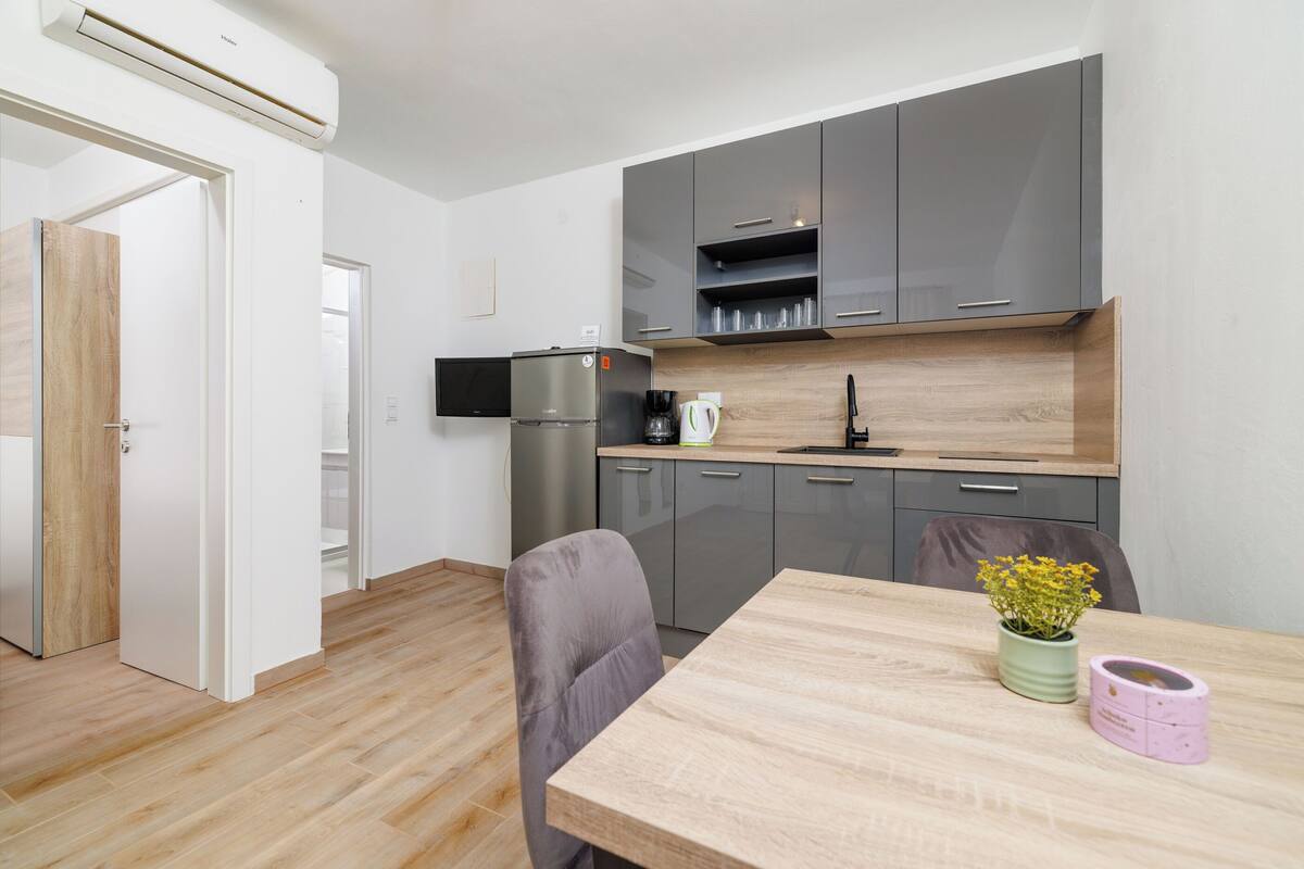 A modern kitchen with grey cabinets, a stainless steel refrigerator, and a wooden dining table with a vase of flowers.