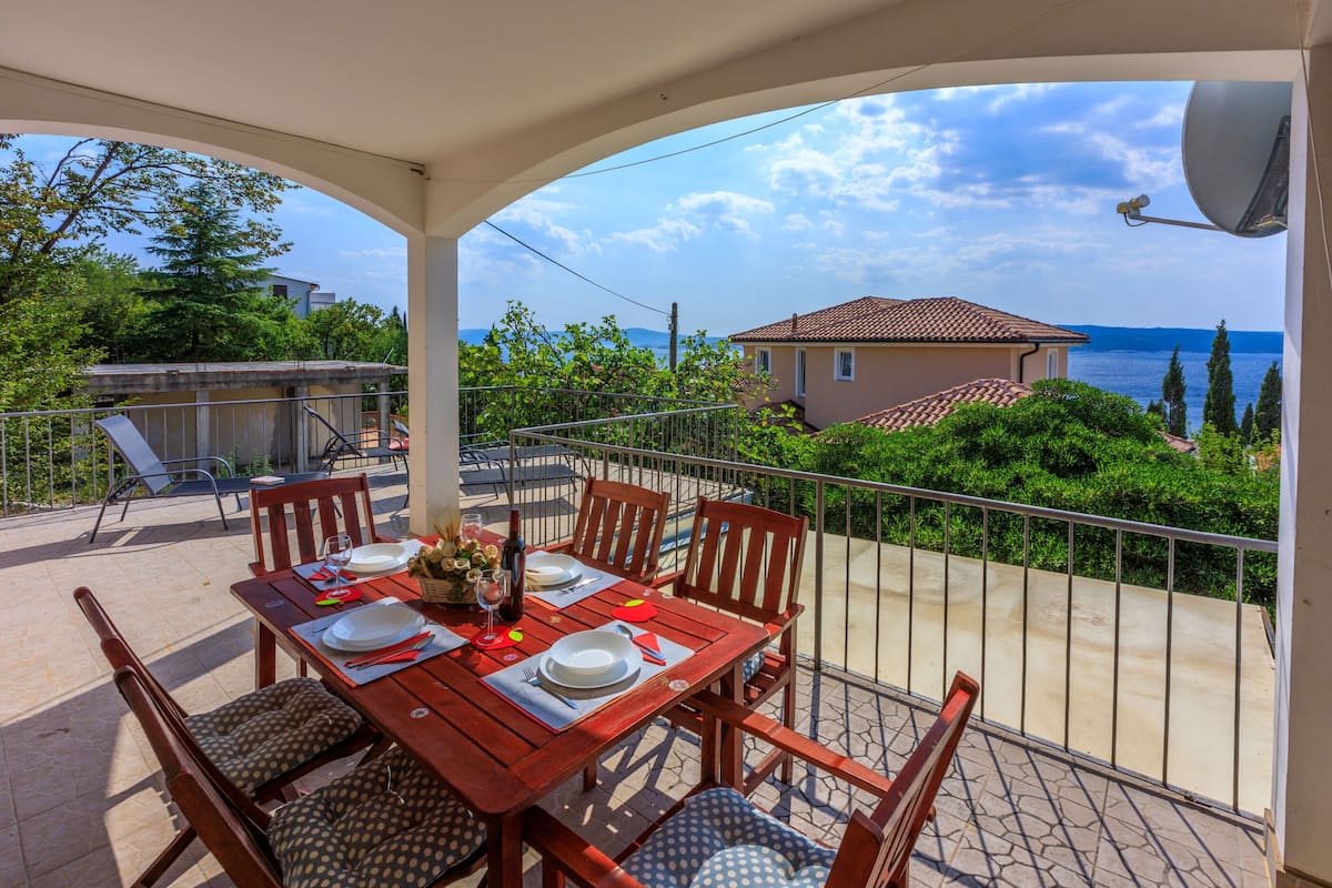Ein Holztisch für vier Personen, Stühle, ein Balkon mit Meerblick und ein Haus im Hintergrund.