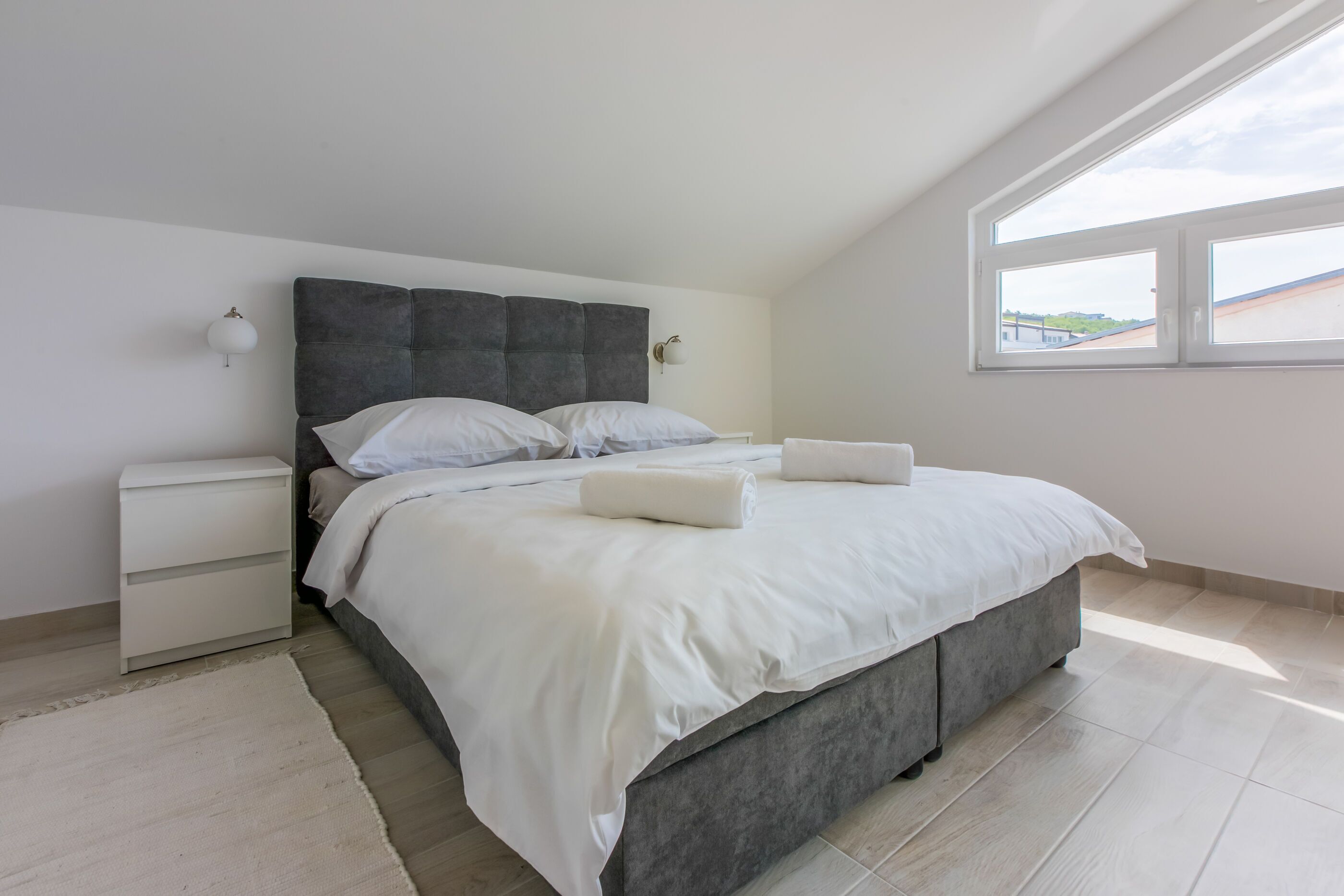 A bedroom with a large bed, a nightstand, a wall-mounted lamp, and a window with a view of buildings.