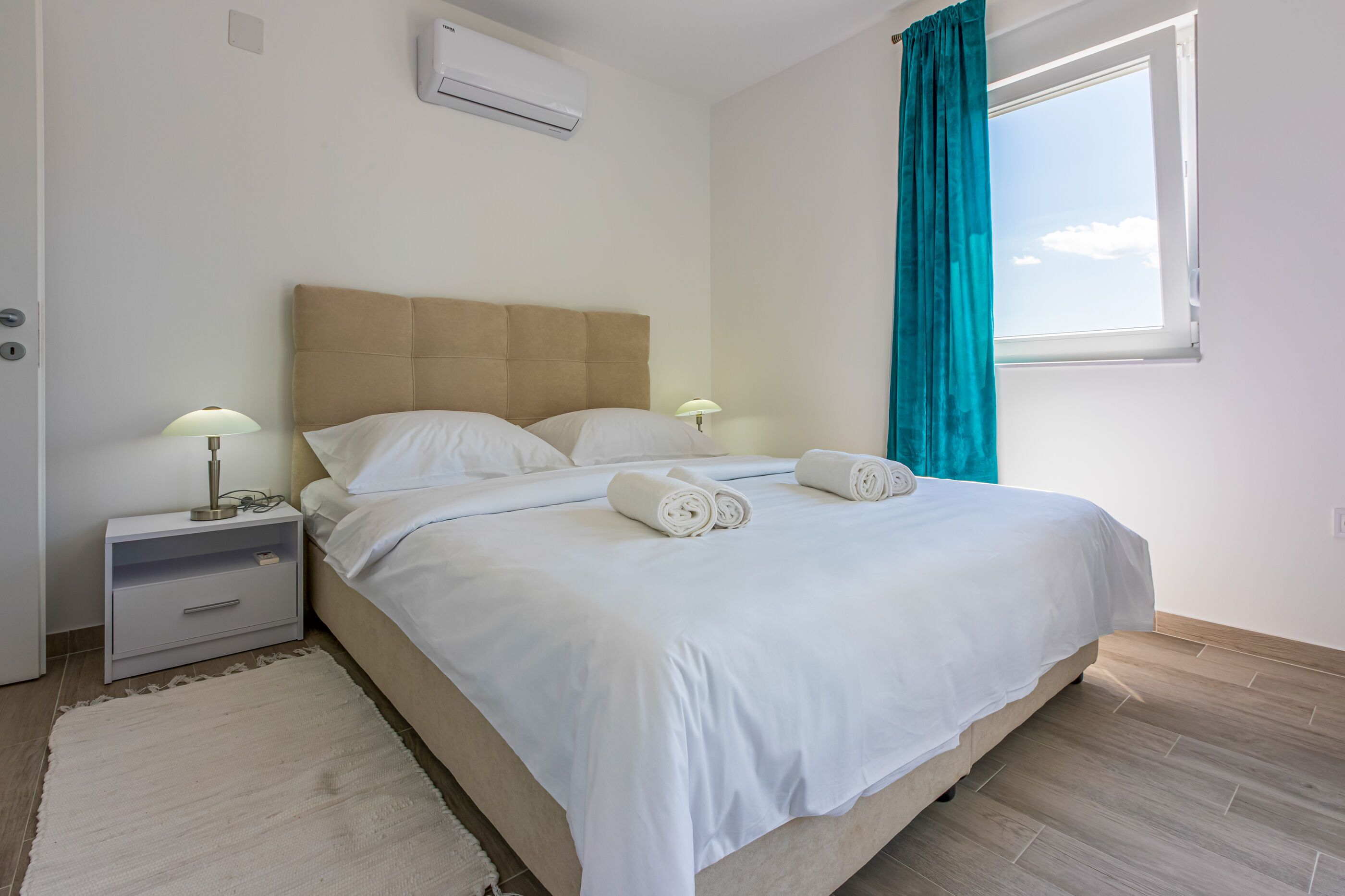 A neatly made bed with white linens and two rolled towels, a bedside table with a lamp, and a window with teal curtains.