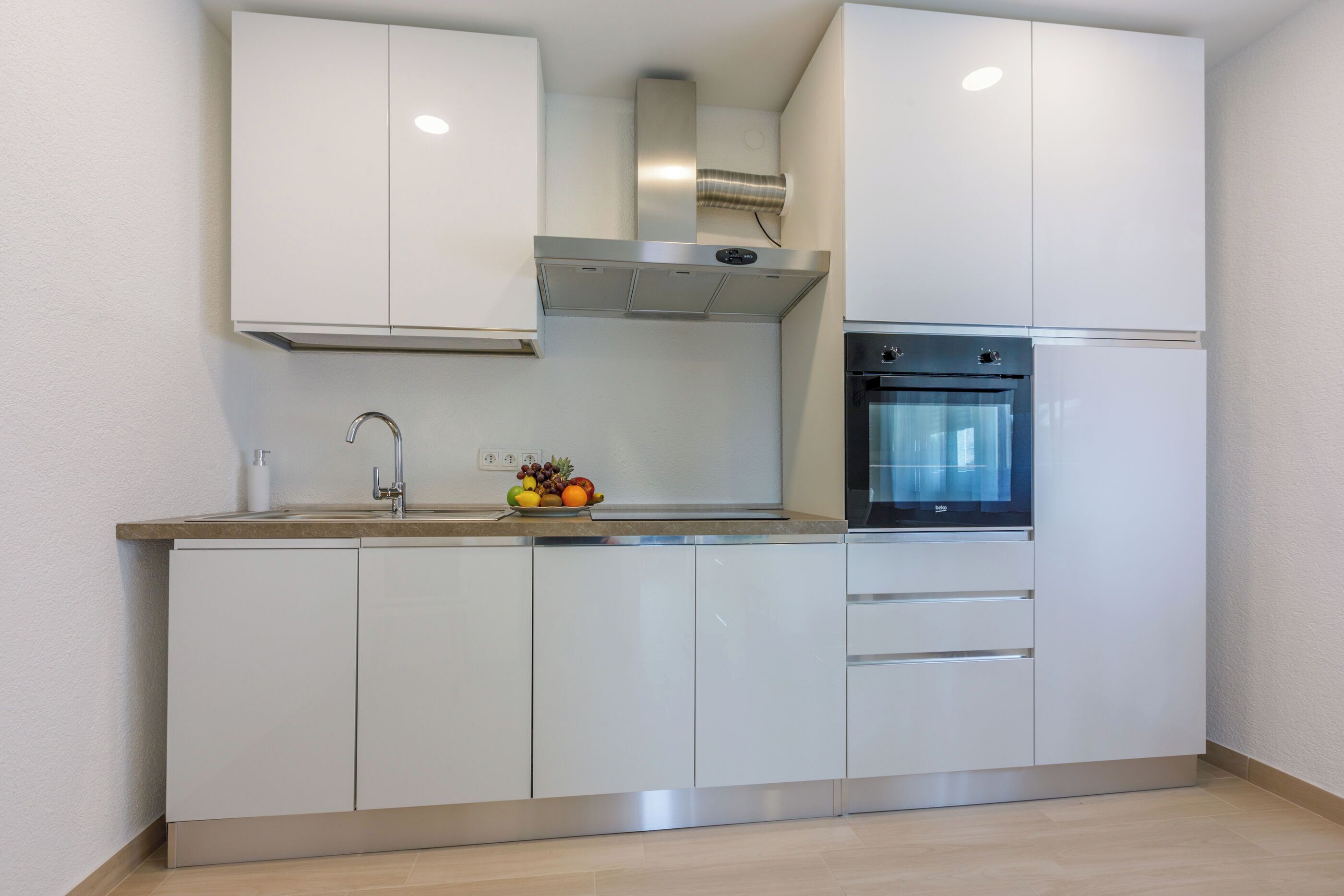 Een moderne keuken met witte kasten, een roestvrijstalen afzuigkap, een inbouwoven en een fruitschaal op het aanrecht.