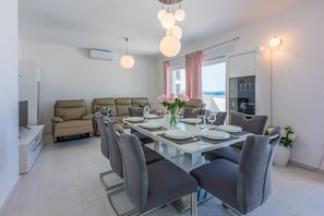A modern living room with a dining area, beige sofas, a flat-screen TV, and a large window with a view. - Apartments Cvetkovic (Crikvenica)