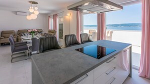 A modern kitchen with a central island, dining area with chairs, and a view of the sea. - Apartments Cvetkovic (Crikvenica)