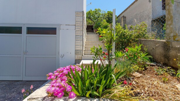 A white building with a gated entrance, a staircase leading to a second floor, and a garden area with pink flowers and green plants. - Apartment Ljubica (Zavalatica)