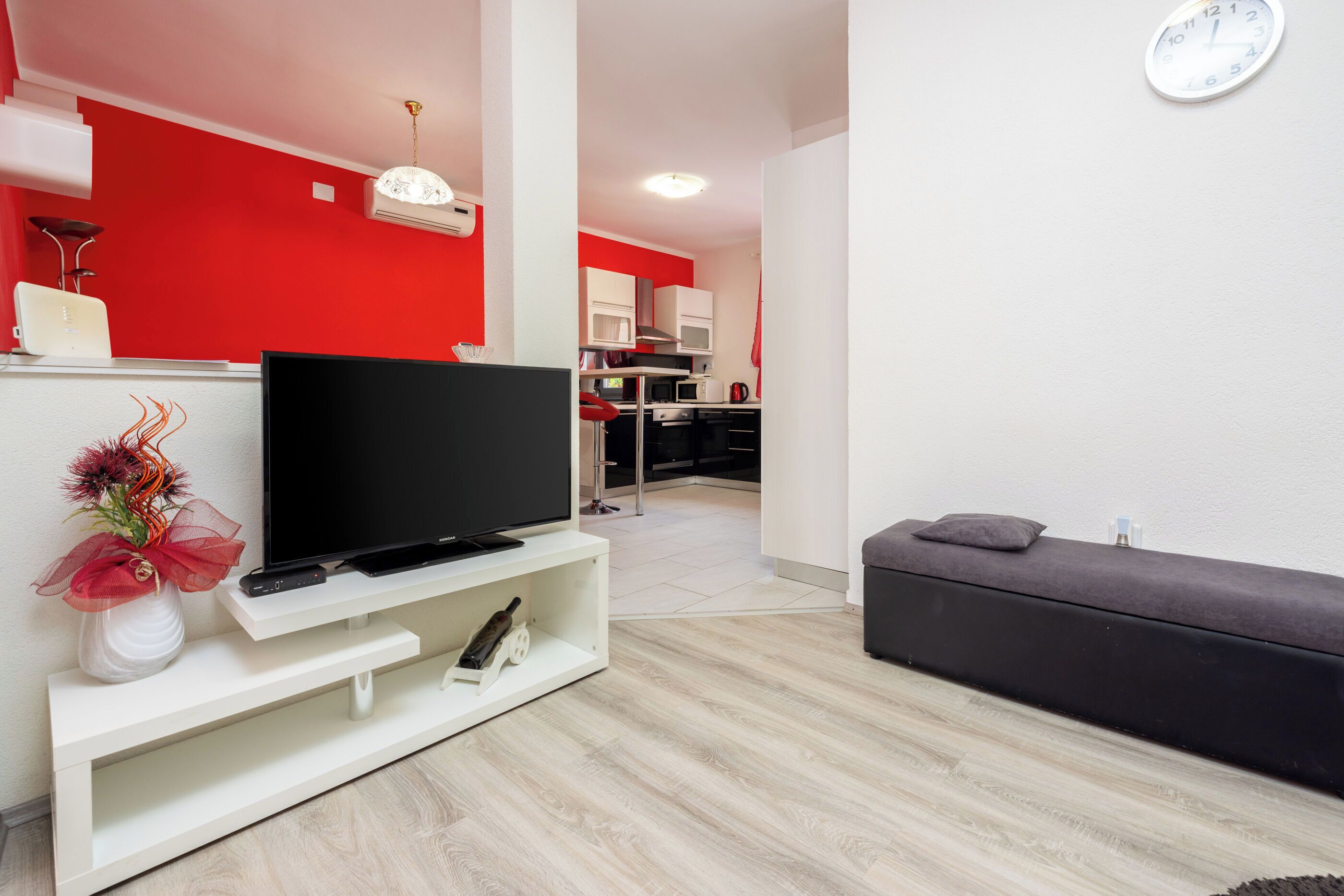 A modern living room with a flat-screen TV on a white stand, a grey sofa, and a red accent wall.