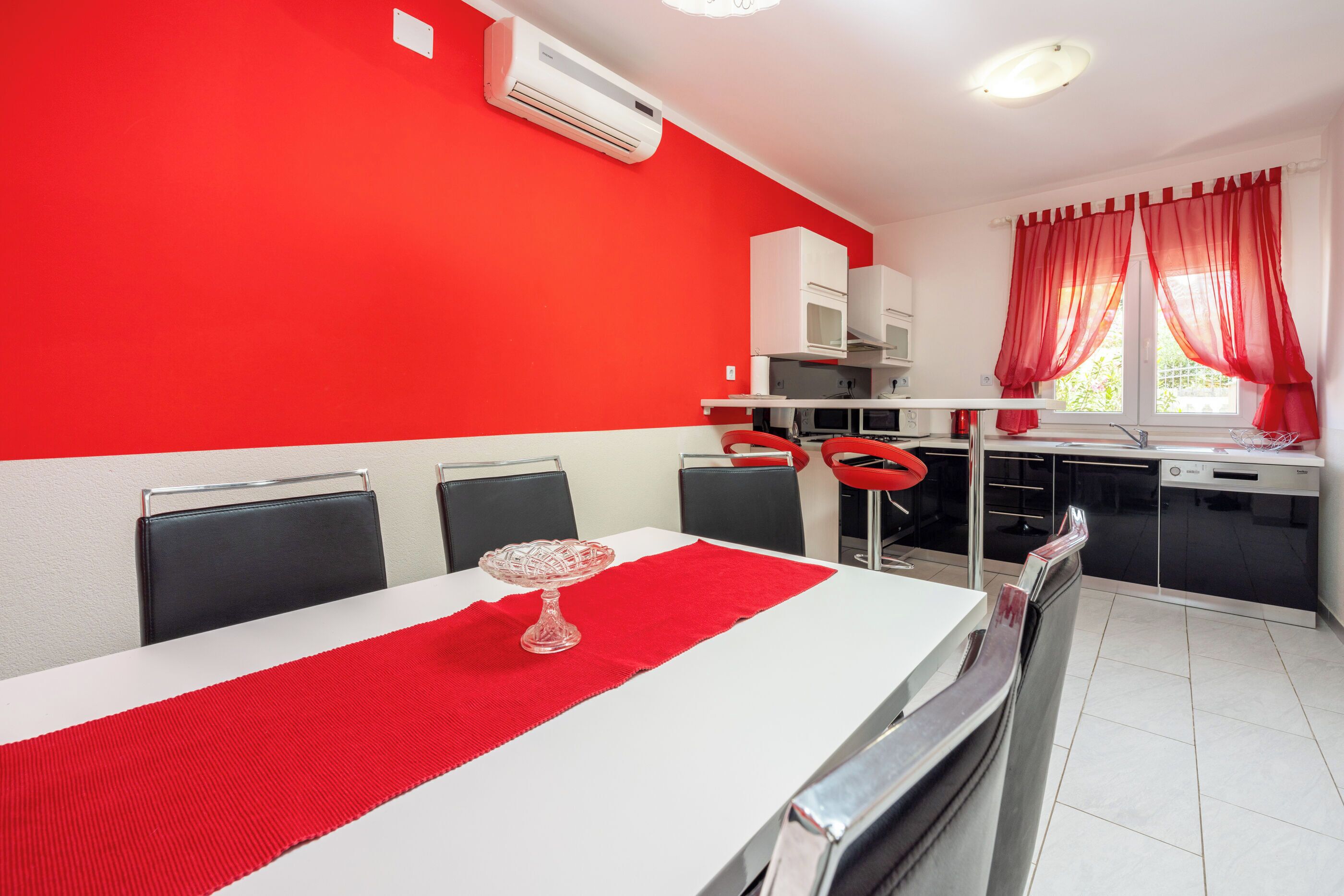 A dining area with a white table, red table runner, and a glass cake plate. There are four black chairs and a white cabinet in the background.