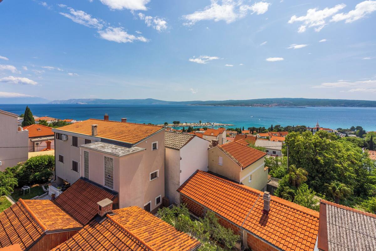 A coastal residential area with terracotta-roofed houses, a clear blue sea, and distant mountains.