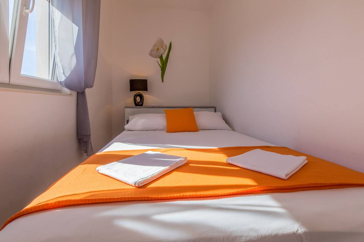 A neatly made bed with an orange bedspread and white pillows, a white flower on the wall, and a small table with a lamp beside the bed.