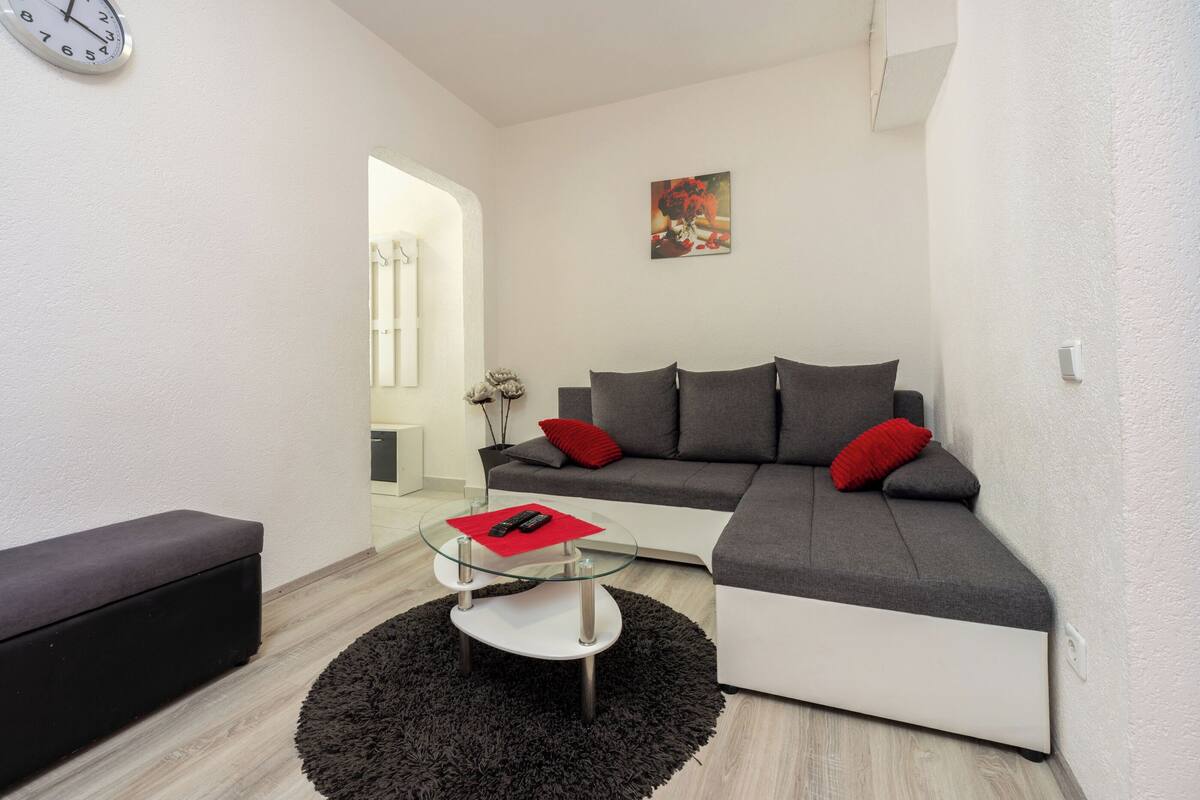A modern living room with a grey sectional sofa, a glass coffee table, a black rug, and a red cushion.