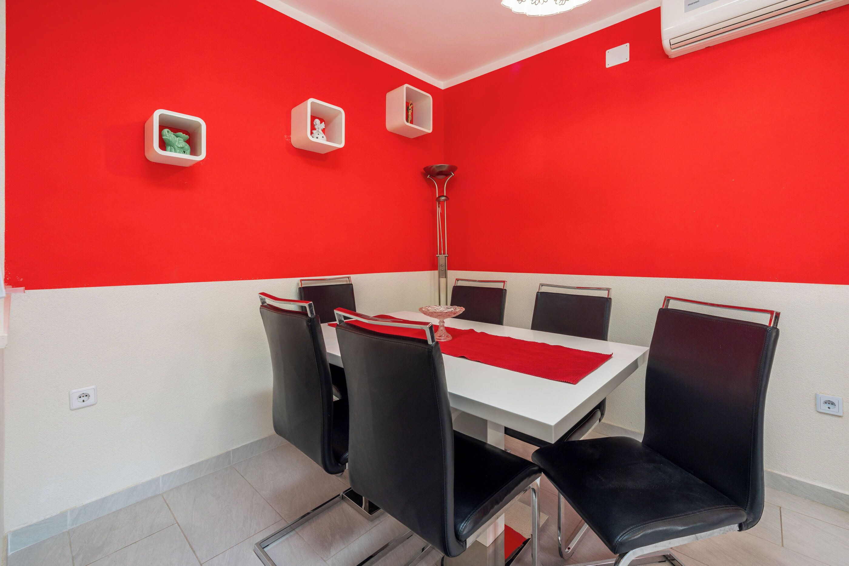 A dining area with a red and white color scheme, featuring a table with chairs, a red runner, and wall-mounted shelves with decorative items.
