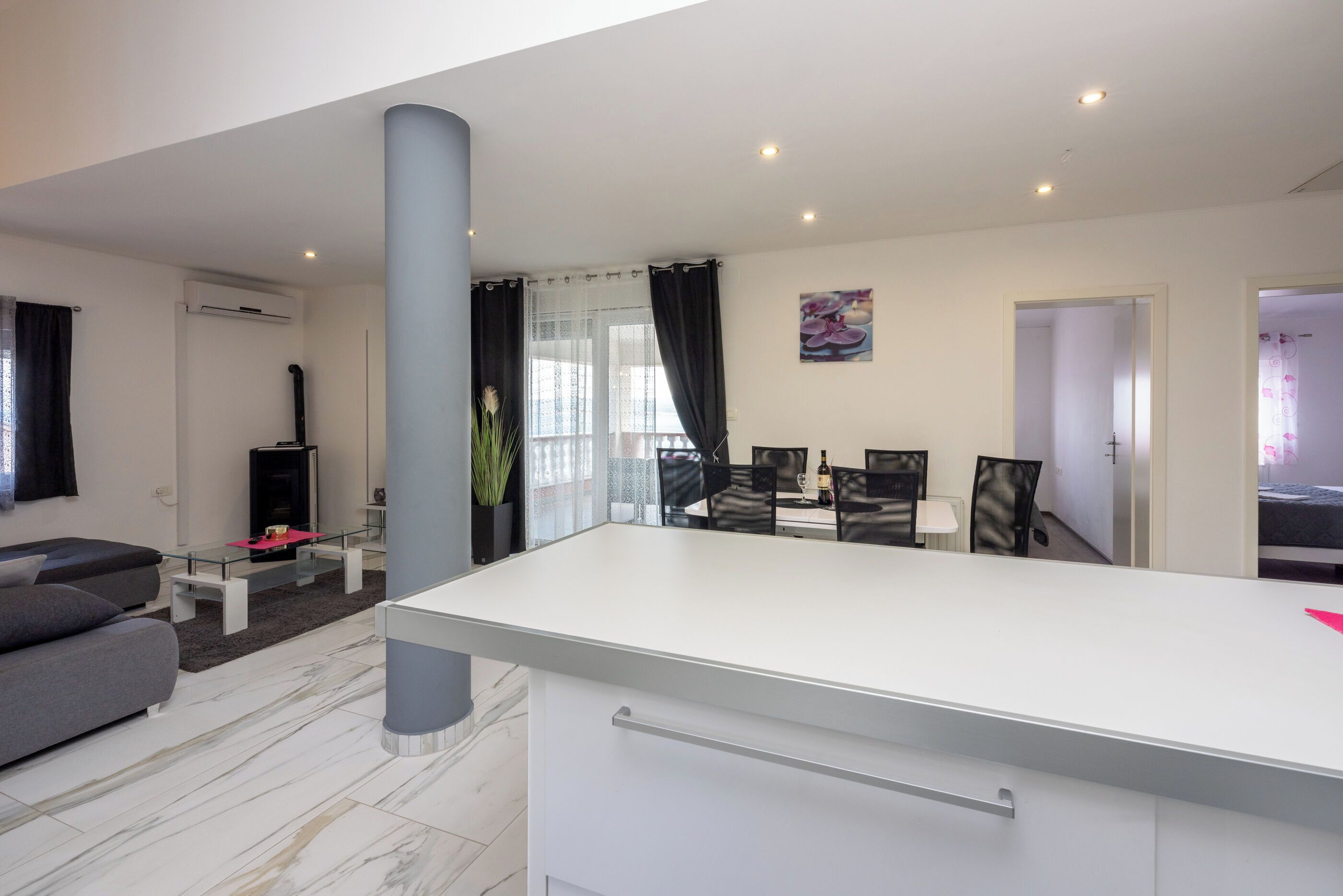 Ein modernes Wohnzimmer mit einem weißen Sofa, einem Glastisch und einem Kamin. Es gibt eine Essecke mit einem weißen Tisch und Stühlen sowie eine Küchenzeile mit weißer Arbeitsplatte und Schränken.