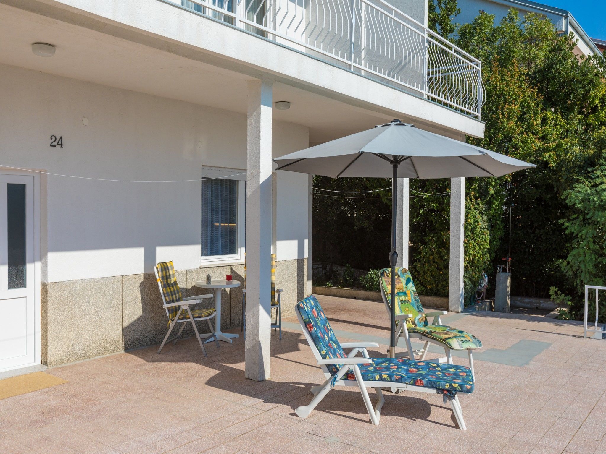 A covered outdoor seating area with two lounge chairs and a table, a white building with the number 24, and a balcony with a white railing.