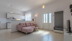 A modern living room with a pink sofa, a small table, and a TV stand. There is a kitchen area with white cabinets and a window with curtains. - Apartment Duda (Novi Vinodolski)
