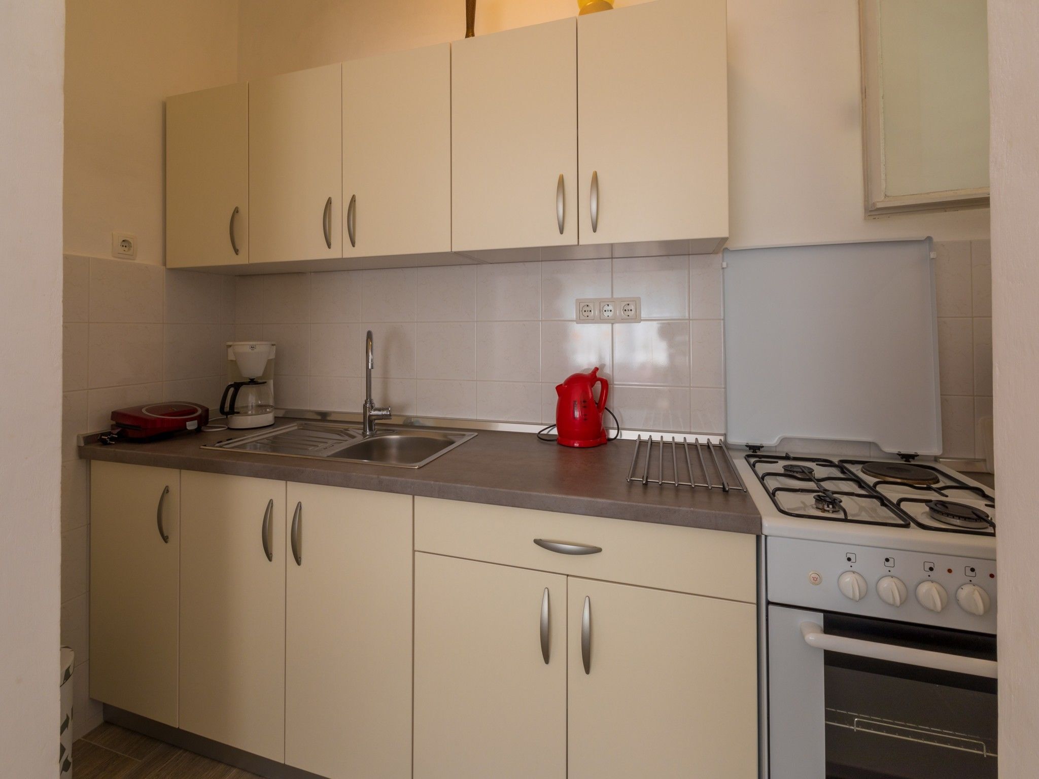 Een keuken met beige kastjes, een roestvrijstalen spoelbak, een rode waterkoker en een gasfornuis.