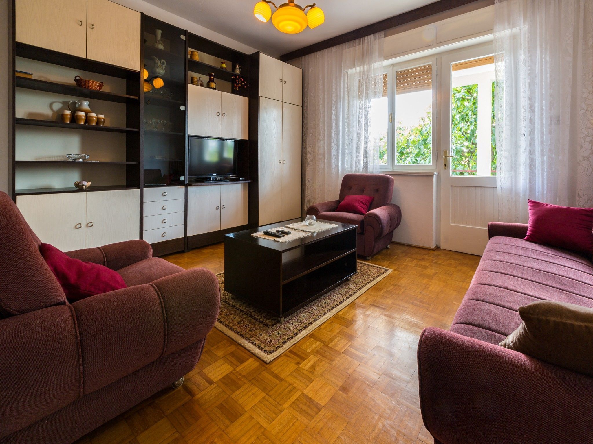 A living room with a sofa, armchair, and a coffee table. There is a television on a cabinet, and a window with curtains.