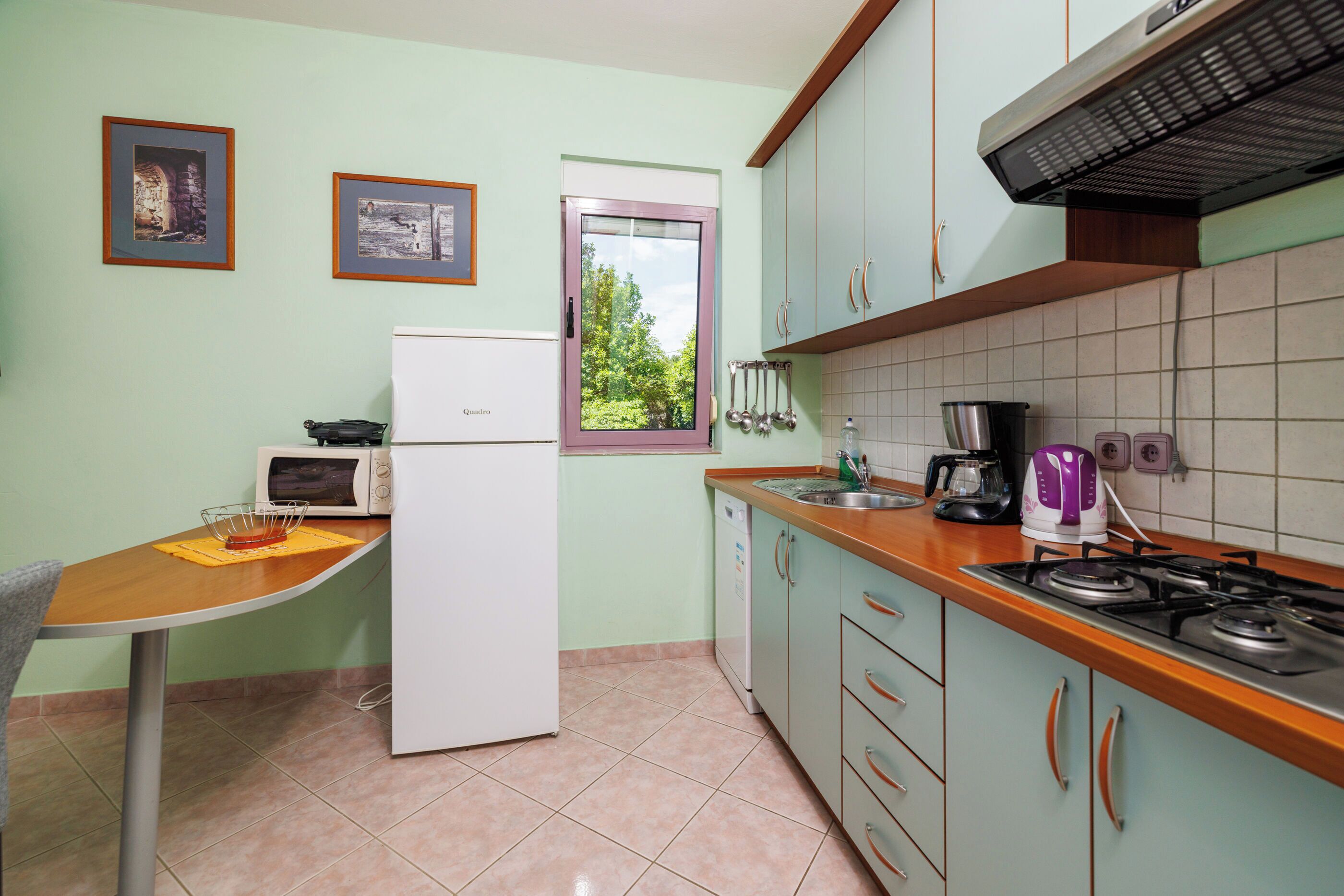 A kitchen with a refrigerator, microwave, sink, and stove. There are cabinets, a window, and two framed pictures on the wall.