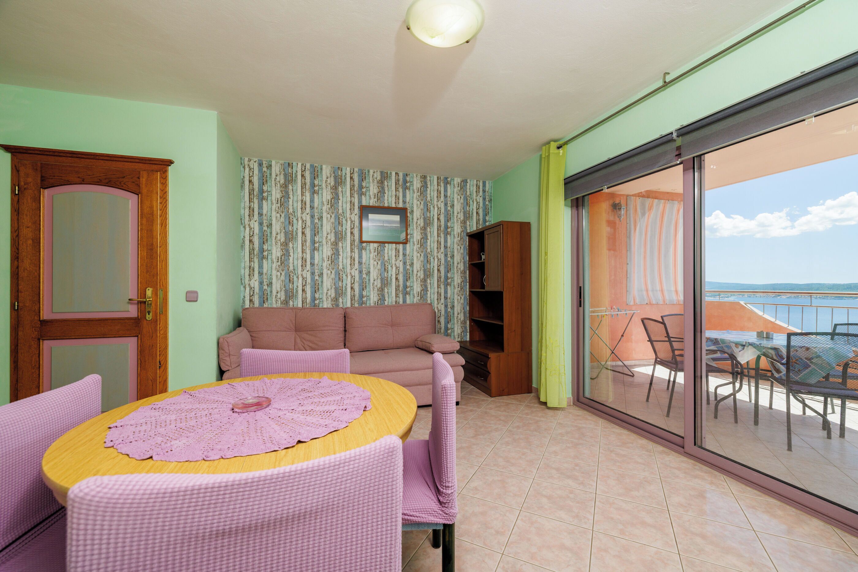 A room with a sofa, a table with a lace tablecloth, a wooden cabinet, and a sliding glass door leading to a balcony with outdoor furniture.