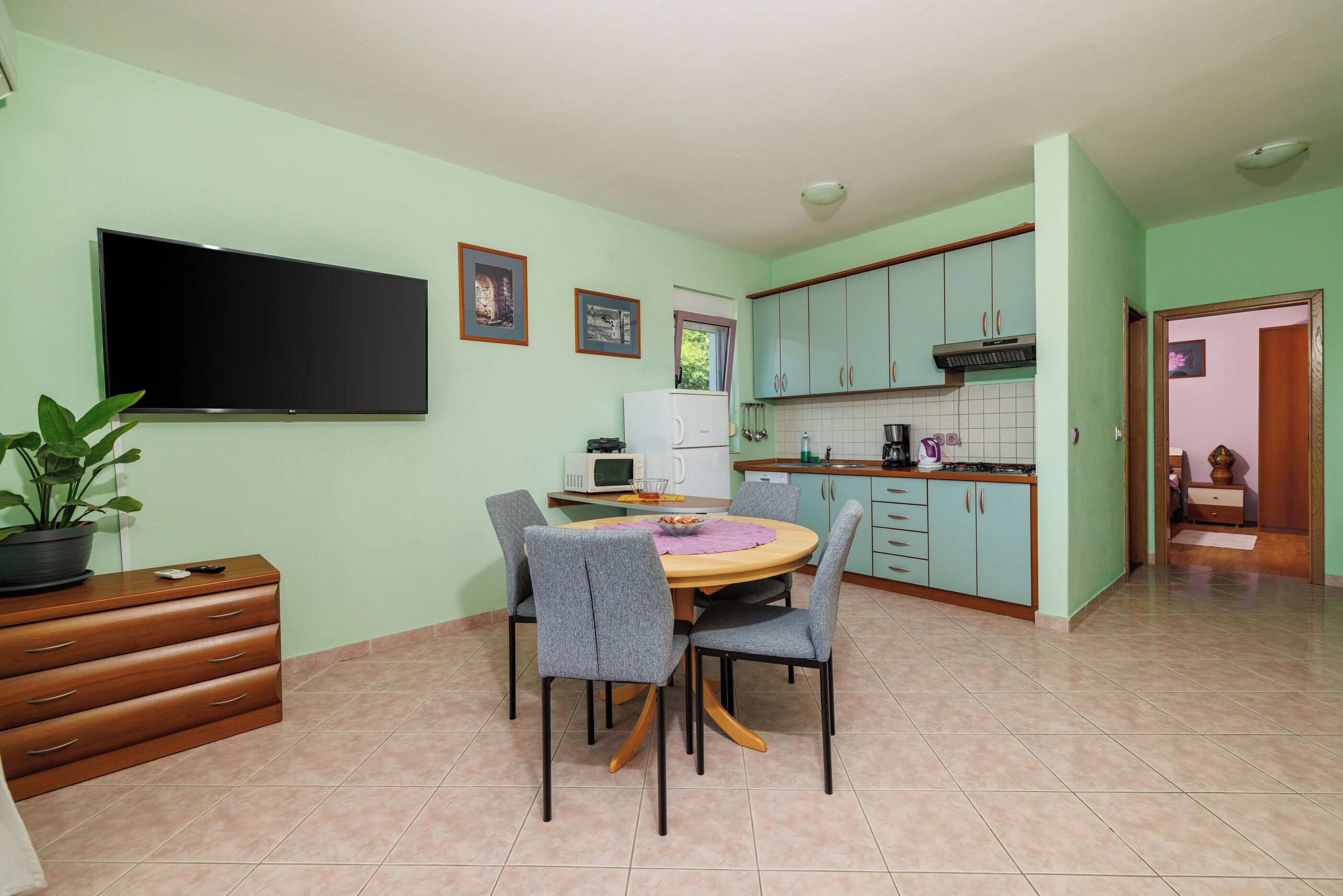 A dining area with a table and chairs, a television, a microwave, and a kitchen area with cabinets and a sink.